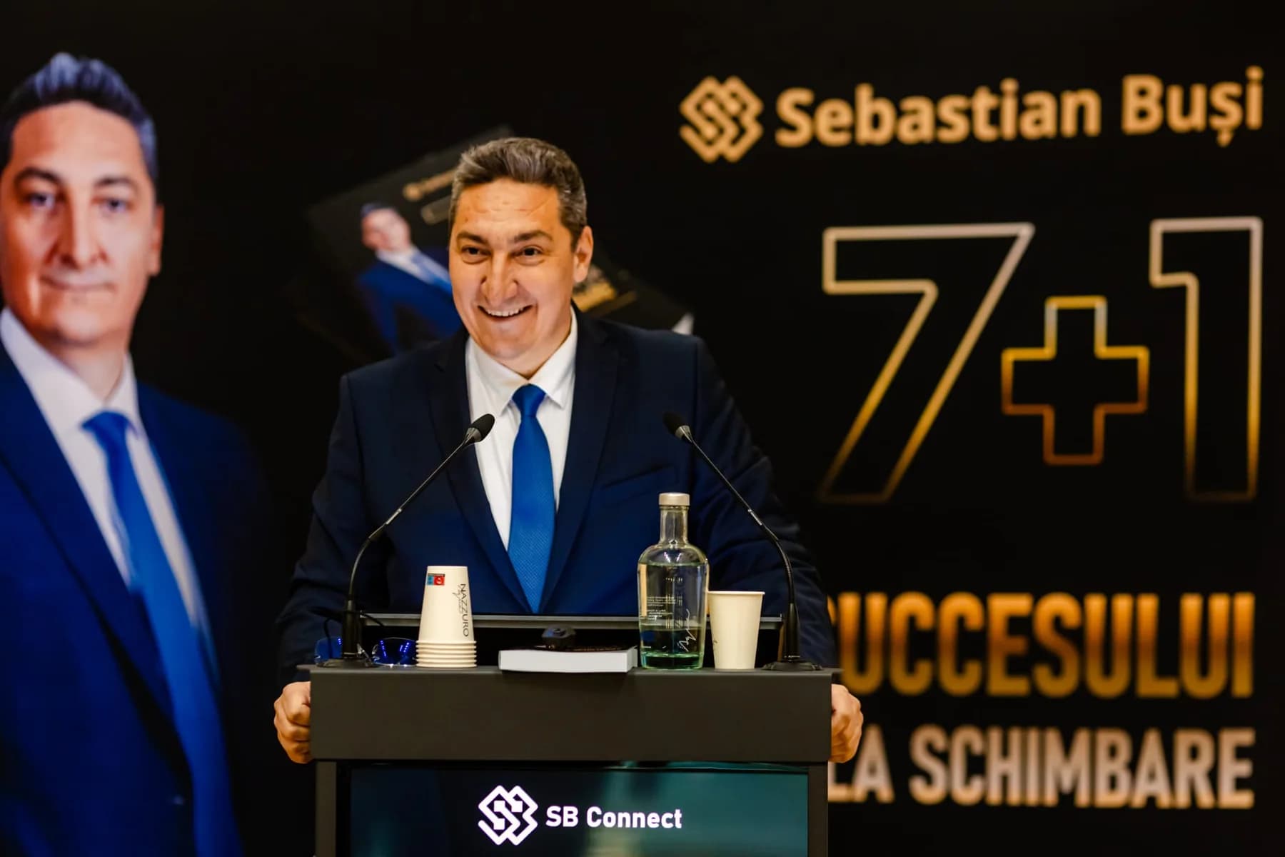 Sebastian Buși vorbind la pupitru, la lansarea cărții 7+1, cu afișul cărții în spate